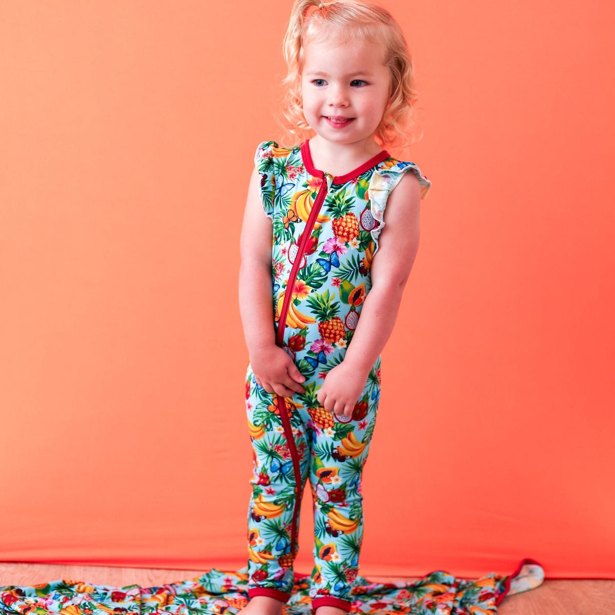 Sleeveless baby romper in a tropical fruit print with dual front zippers, worn by a smiling child.