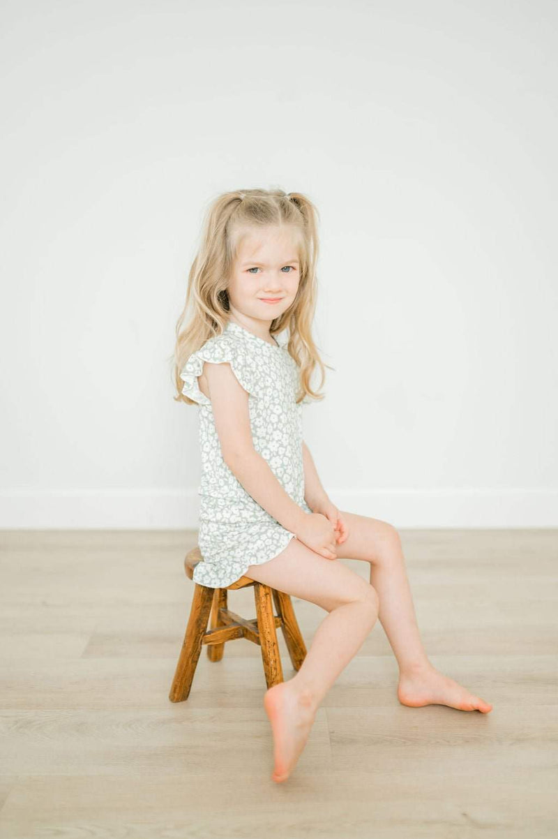 Girl wearing a sage ditsy floral two-piece short set with ruffle details sitting on a stool.
