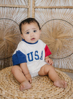 Baby wearing a red, white, and blue colorblock t-shirt romper from Little Joy Co., playing happily.