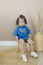 Little Joy Co. 100% cotton baby t-shirt romper in a neutral color, laid flat on a soft surface.